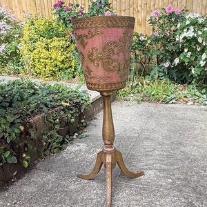 May include: A vintage pink and gold plant stand. The planter features a pink fabric exterior with gold scroll detailing. The wooden stand has a tripod base. The stand is outside, surrounded by plants and flowers.