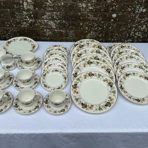 May include: A full vintage dinner service set, comprising cream-coloured plates, saucers, cups, and serving dishes. The set includes a serving platter, a creamer, and a sugar bowl. Each piece is decorated with a leaf pattern in brown and green.