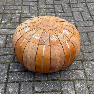 May include: A round, tan leather Moroccan pouf with geometric and floral patterns. The pouf has silver metallic accents and a stitched design. The top features a circular design with a central emblem. The pouf is made of leather and has a soft, rounded shape.