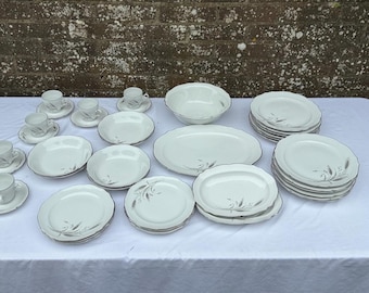 Op de afbeelding: Een complete witte porseleinen servies set met een zilveren rand en een delicaat bloemmotief. De set bevat kopjes, schotels, kommen, borden en serveerschalen, allemaal gerangschikt op een wit tafelkleed.