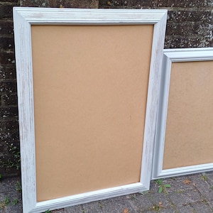 May include: Two white picture frames with a distressed finish. The larger frame is standing upright and the smaller frame is leaning against it. Both frames are empty.