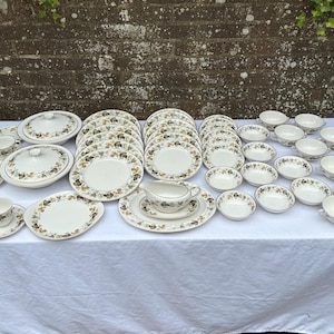 May include: A full vintage cream china dinner service with a floral design. The set includes plates, bowls, cups, saucers, serving dishes, and a creamer. The items are arranged on a white tablecloth.