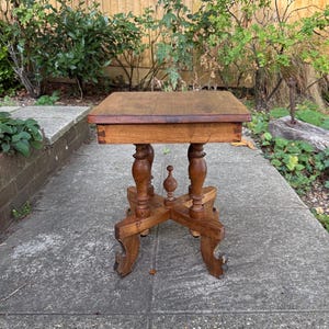 May include: A small square wooden side table with a smooth top surface and visible dovetail joints on the apron. The table features four turned legs connected by a cross-shaped base with a central decorative finial and curved feet.
