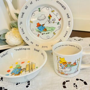 May include: A white ceramic children's dish set featuring Paddington Bear illustrations. The set includes a plate, bowl, and mug, each with colorful drawings of Paddington in various scenes. Text is included on each piece.