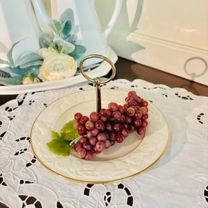 May include: A two-tiered serving stand with a gold handle. The bottom tier is a white plate with a gold rim, holding a cluster of red grapes and a green leaf. The stand sits on a white lace doily.