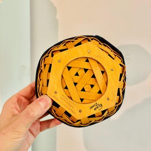 May include: A small, round, woven basket with a black rim and a light brown wooden base. The base features a geometric design and the text "LONGABERGER BASKETS HANDWOVEN DESIN, OHIO USA". The basket is held by a hand.
