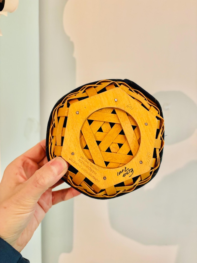 May include: A small, round, woven basket with a black rim and a light brown wooden base. The base features a geometric design and the text "LONGABERGER BASKETS HANDWOVEN DESIN, OHIO USA". The basket is held by a hand.