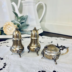 May include: A set of antique silver-colored serving pieces, including two salt shakers and a small lidded container. The pieces have ornate detailing and stand on small, decorative feet. The set is displayed on a white, patterned tablecloth.