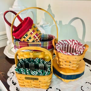 May include: A collection of woven baskets in various sizes and shapes. The baskets feature wooden handles and colorful fabric liners in red, green, and plaid patterns. One basket has a Longaberger label.