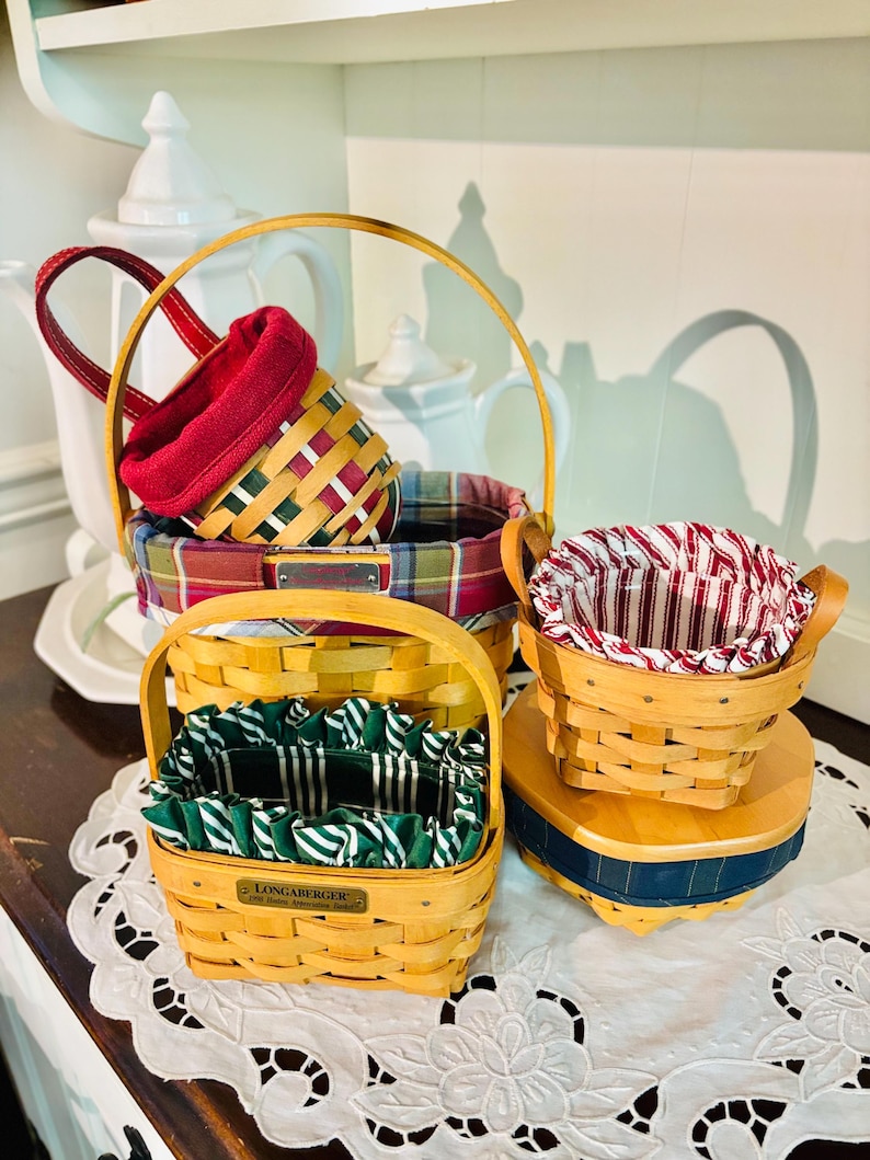 May include: A collection of woven baskets in various sizes and shapes. The baskets feature wooden handles and colorful fabric liners in red, green, and plaid patterns. One basket has a Longaberger label.