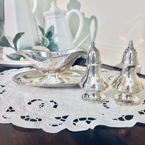 May include: A silver gravy boat with a matching tray and two silver salt and pepper shakers. The items are on a white lace doily. White teapots and a creamer are in the background.