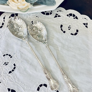May include: A pair of silver-toned metal salad serving spoons featuring intricate embossed fruit and leaf patterns on the bowls and decorative shell-like motifs on the handles. The utensils have a polished finish and long, slender stems.