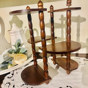 May include: Two small, dark brown wooden tiered display stands. Each stand has a half-circle base, a middle shelf, and a top shelf. The stands are made of turned wood with a glossy finish. The stands are decorative and suitable for displaying small items.