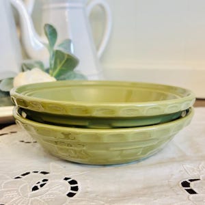 May include: Two light green ceramic bowls stacked on top of each other. The bowls have a textured, basket-weave design around the sides and a scalloped rim. The bowls are sitting on a white, embroidered tablecloth.
