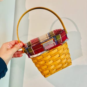 May include: A yellow woven basket with a wooden handle and a plaid fabric lining. The basket has a rectangular shape and a label on the front. The handle is arched and made of wood.