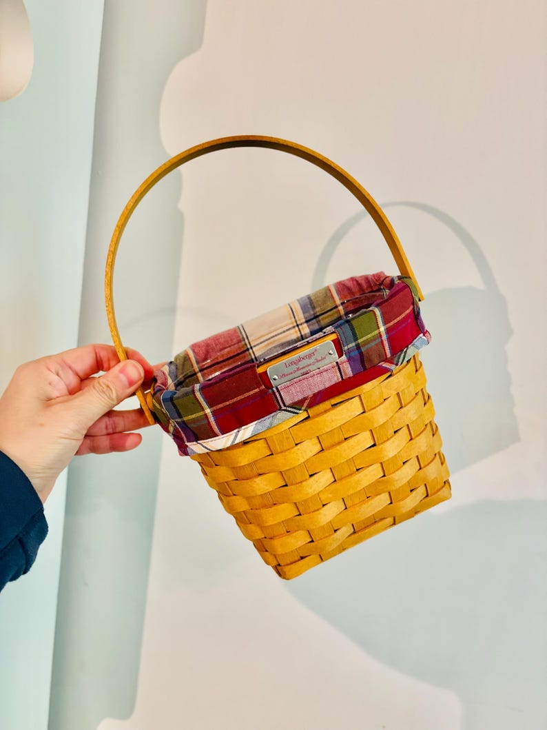 May include: A yellow woven basket with a wooden handle and a plaid fabric lining. The basket has a rectangular shape and a label on the front. The handle is arched and made of wood.
