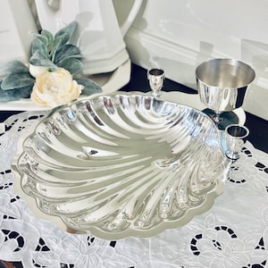 May include: A polished silver-toned serving dish shaped like a large scallop shell with a scalloped edge. The dish features a reflective, smooth surface with radiating fluted grooves. It is presented alongside a small silver-toned cup.