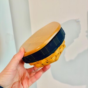 May include: A small, round basket with a wooden lid. The basket is woven in a light brown color, with a navy blue fabric band around the middle. The lid is made of light-colored wood.