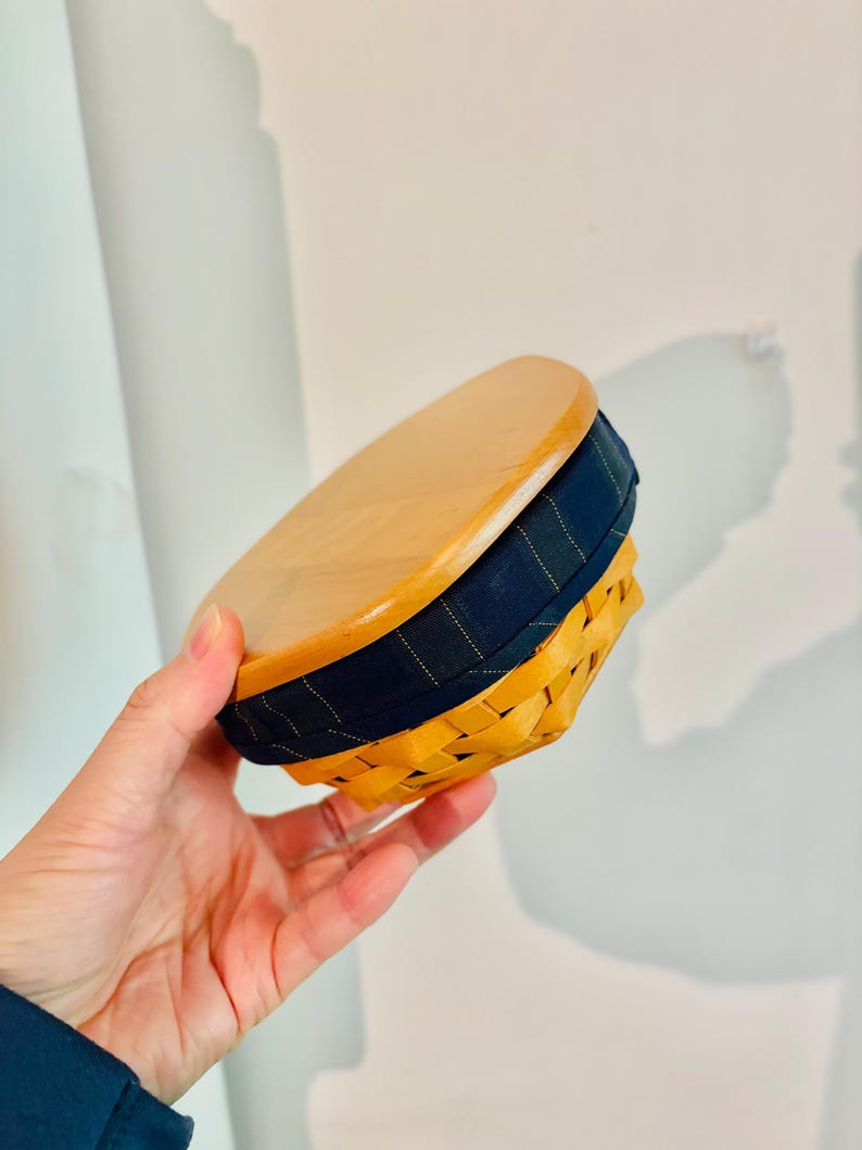 May include: A small, round basket with a wooden lid. The basket is woven in a light brown color, with a navy blue fabric band around the middle. The lid is made of light-colored wood.