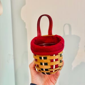 May include: A small, woven basket with a red fabric lining and a matching red handle. The basket features a pattern of tan, black, and red woven strips. The handle is made of red leather-like material. The basket is held in a person's hand.