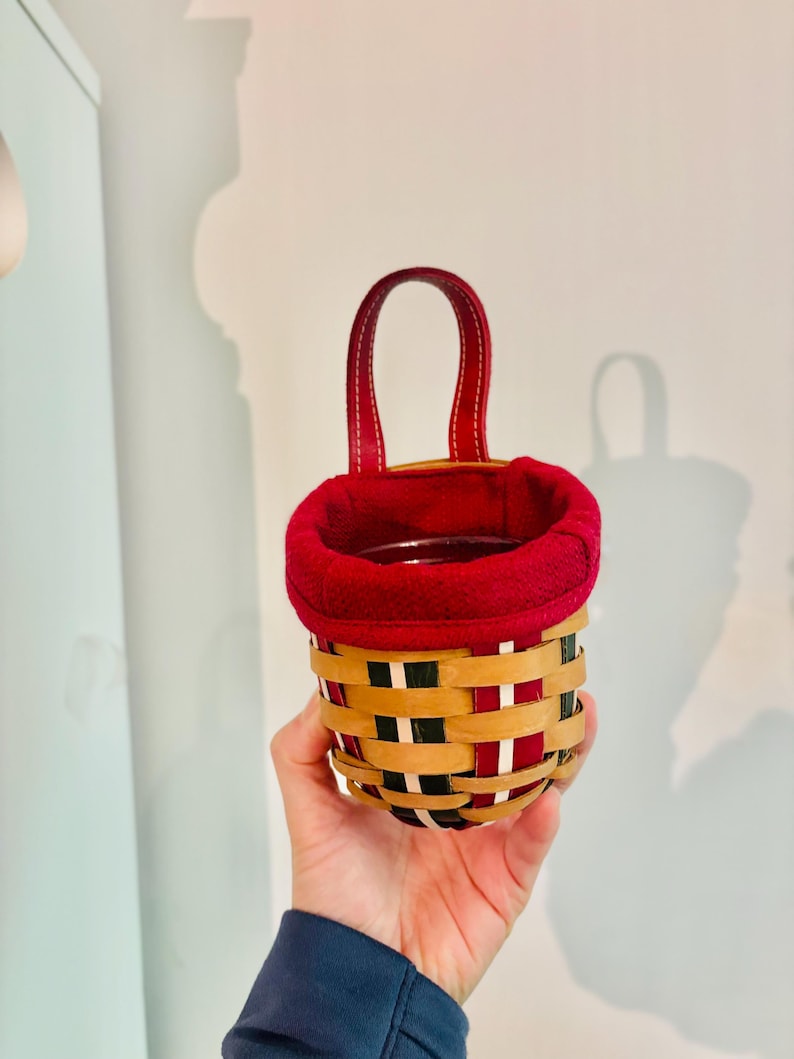May include: A small, woven basket with a red fabric lining and a matching red handle. The basket features a pattern of tan, black, and red woven strips. The handle is made of red leather-like material. The basket is held in a person's hand.