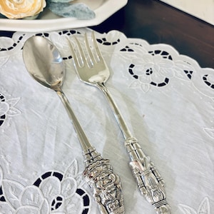 May include: Silver spoon and fork set with ornate handles. The spoon has a rounded bowl, and the fork has four tines. The handles feature detailed, sculpted designs. The set is displayed on a white, embroidered tablecloth.