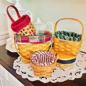 May include: A collection of woven baskets with fabric liners in various sizes and patterns. The baskets feature red, green, and plaid fabrics. One basket has a handle, and another is on a wooden stand. The baskets are on a white lace doily.