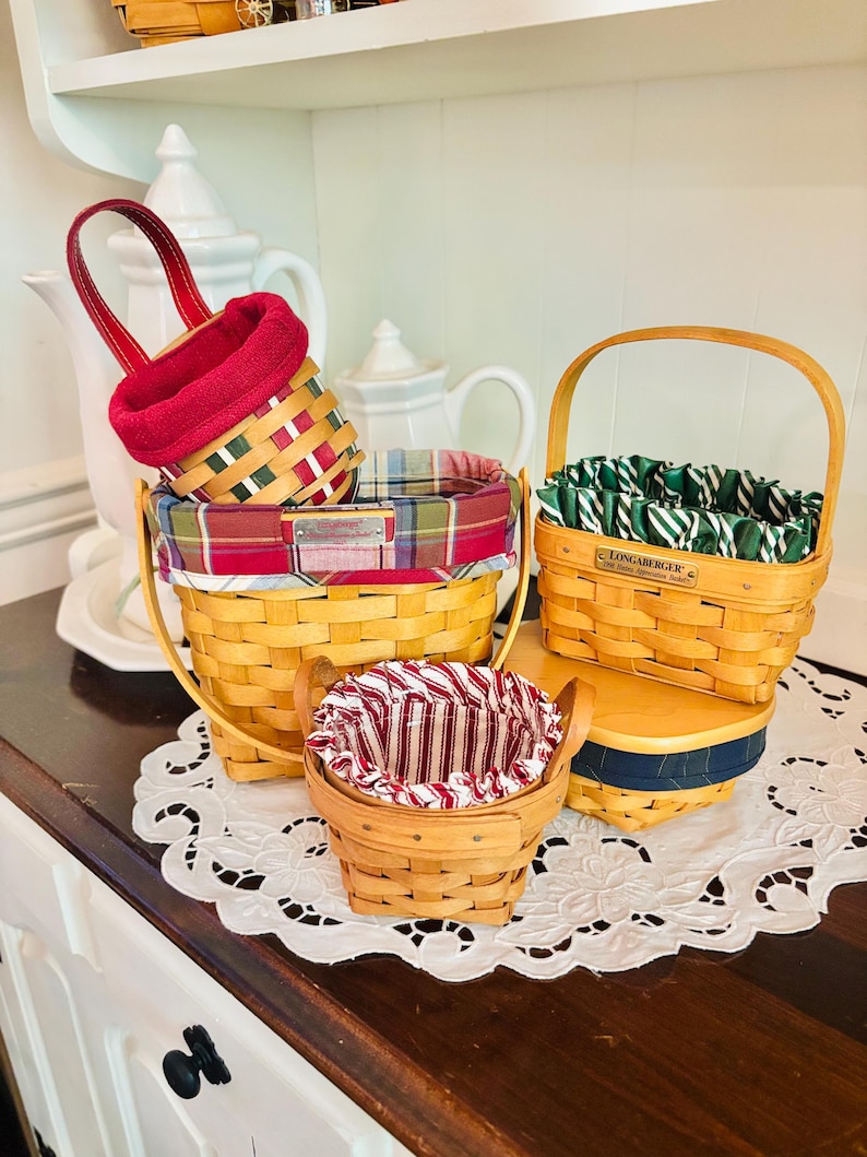 May include: A collection of woven baskets with fabric liners in various sizes and patterns. The baskets feature red, green, and plaid fabrics. One basket has a handle, and another is on a wooden stand. The baskets are on a white lace doily.