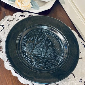 May include: A round ceramic plate with a wide, dark charcoal rim and a textured, mottled blue and black center featuring a stylized tree-like pattern. The plate has a glossy finish and a distinct, raised border separating the rim from the center.
