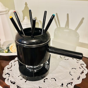 May include: A black fondue pot with a handle and a set of fondue forks with colored tips. The pot sits on a black base, and the forks are arranged inside. The pot is on a white lace doily.