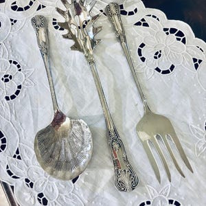 May include: Three antique silver serving utensils: a shell-shaped spoon, a spaghetti server, and a fork. The utensils have ornate handles with a leaf design. They are arranged on a white, embroidered doily.