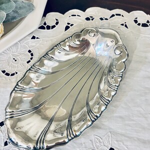 May include: A polished silver-toned metal dish shaped like a seashell with a scalloped edge and embossed radial lines extending from the base to the rim. The dish has a reflective, smooth surface and an elongated oval form with decorative curves.