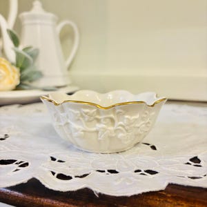 May include: A small, white, floral-shaped bowl with a gold rim. The bowl has a detailed embossed design of vines and leaves. It sits on a white, lace-edged doily. Other white ceramic pieces are in the background.