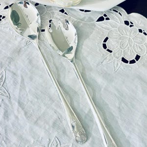 May include: Two silver serving spoons with ornate floral designs on the handles rest on a white, embroidered tablecloth. A white plate with a yellow flower and green leaves is in the background. The spoons are reflective.