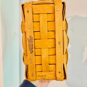 May include: A rectangular, light brown woven basket with vertical and horizontal wooden strips. The basket has a handle and is secured with metal rivets. The text "Longaberger Handmade Baskets" is visible on the side.