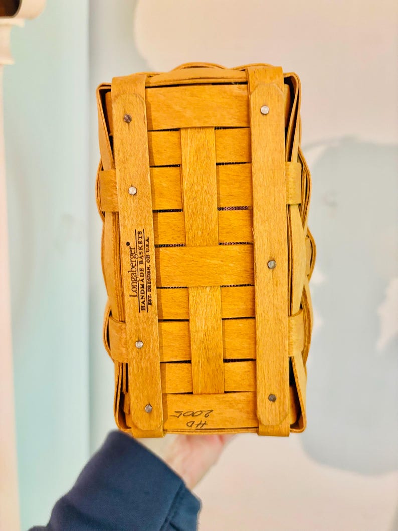 May include: A rectangular, light brown woven basket with vertical and horizontal wooden strips. The basket has a handle and is secured with metal rivets. The text "Longaberger Handmade Baskets" is visible on the side.