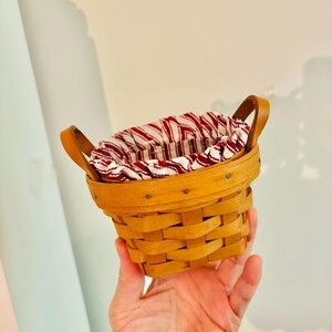 May include: A small, round, woven basket with a light brown finish. It features two arched leather handles and a red and white striped fabric lining the interior. The basket is designed for storage or display.