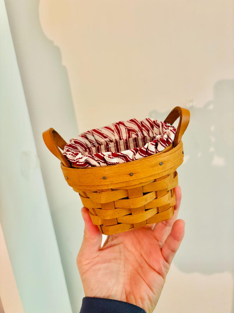May include: A small, round, woven basket with a light brown finish. It features two arched leather handles and a red and white striped fabric lining the interior. The basket is designed for storage or display.