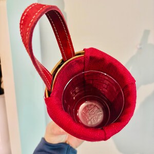 May include: A red suede cup holder with a stitched handle. The holder is designed to fit a glass, visible inside. The holder has a wooden rim and is held by a person.