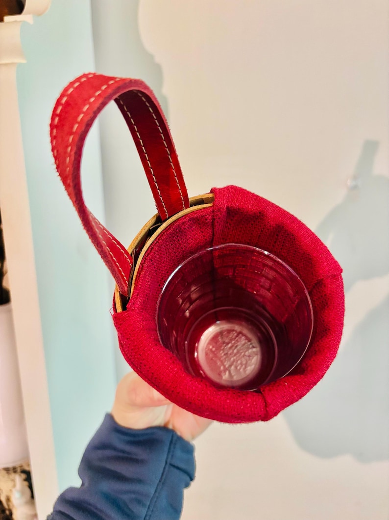 May include: A red suede cup holder with a stitched handle. The holder is designed to fit a glass, visible inside. The holder has a wooden rim and is held by a person.