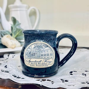 May include: A dark blue ceramic mug with a rounded body and a handle. The front features a cream-colored oval decal with a line drawing of a historic building, the year 1760, and the text Speedwell Forge Bed & Breakfast, Lititz, Pennsylvania.