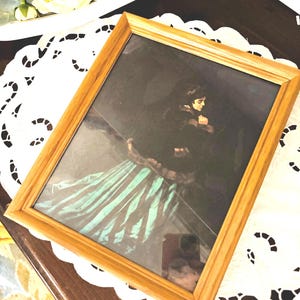 May include: A framed print featuring a woman in a long dress with teal and white stripes. The frame is made of light-colored wood. The image is set on a white lace doily.