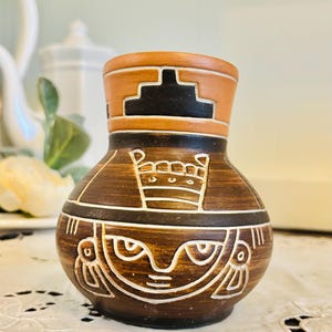 May include: A small, brown ceramic vase with a unique design. The vase features a detailed pattern of white and black geometric shapes and figures. The top rim is a lighter brown color. The vase is likely a decorative item.
