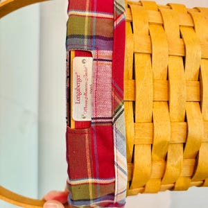 May include: A woven basket with a wooden handle and a plaid fabric lining. The fabric is red, green, and blue. A metal tag reads "Longaberger" and "Women Memories Basket 2005".