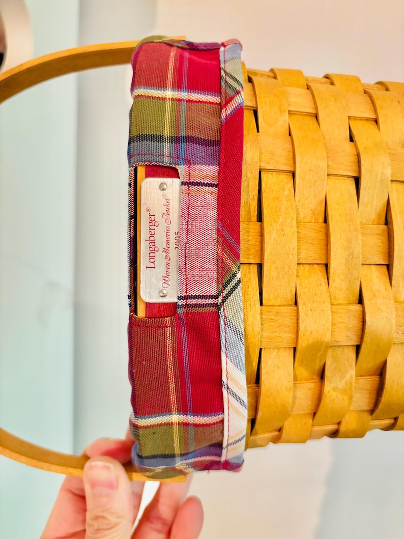May include: A woven basket with a wooden handle and a plaid fabric lining. The fabric is red, green, and blue. A metal tag reads "Longaberger" and "Women Memories Basket 2005".