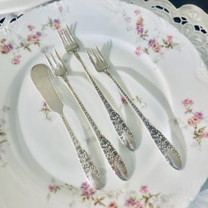 May include: Silverware set including three forks and a butter knife, arranged on a floral patterned plate. The silverware features a detailed floral design on the handles. The plate has a white background with pink and purple floral accents.