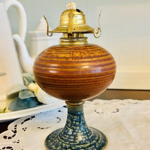May include: A vintage oil lamp with a brown, striped ceramic body and a brass top. The base is a textured blue ceramic. The lamp has a wick and is designed for oil. Decorative lighting.