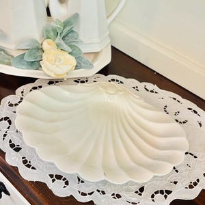 May include: A white ceramic serving dish shaped like a large scallop shell with a glossy finish and radial ridges. The dish is centered on a white oval lace doily featuring intricate floral cutwork patterns and scalloped edges.