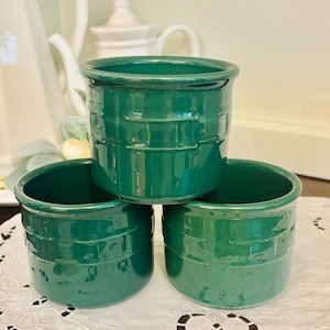 May include: Three stacked, emerald green ceramic ramekins. Each ramekin has a textured, basketweave-like design around the exterior. The ramekins are sitting on a white, lace-edged doily. Teapots are visible in the background.
