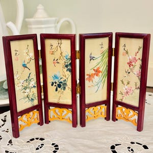 May include: A miniature folding screen with four panels. Each panel features a painting of birds and flowers in vibrant colors. The frames are a deep red, and the base is a decorative gold color.